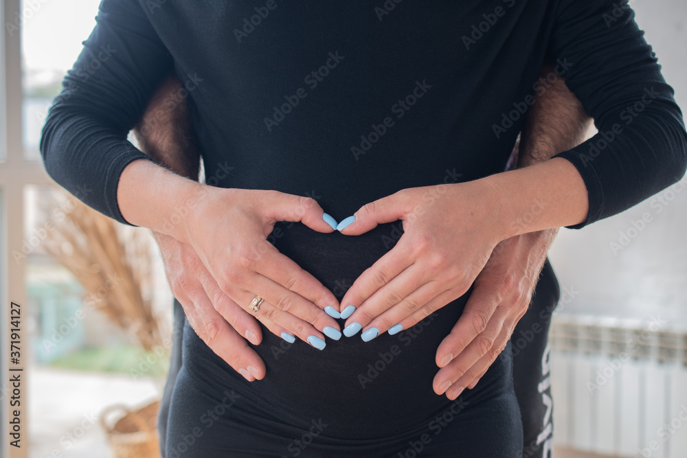 A pregnant woman shows a heart with her hands on her stomach and men's hands help to hold the tummy where the baby is: the concept of women's health and full families A pregnant woman shows a heart with her hands on her stomach and men's hands help to hold the tummy where the baby is: the concept of women's health and full families