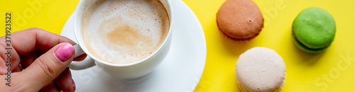 Woman hand holds a cup of coffee, cake macaron on yellow table from above. Female working desk. Cozy breakfast.white cup of tasty coffee in a woman hand and colorful macaroons. Banner size.