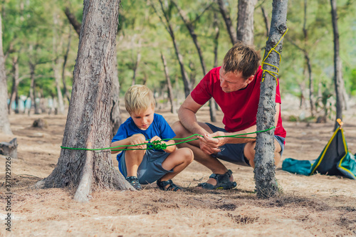 Camping people outdoor lifestyle tourists in summer forest near lazur sea. Blond boy son with father study survival techniques, practice methods of tying rope knots. Natural children education