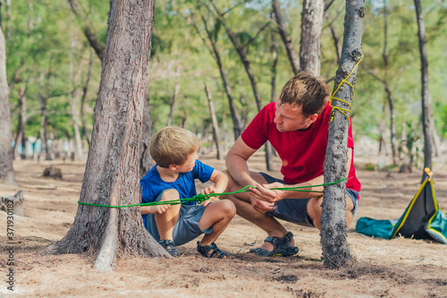Camping people outdoor lifestyle tourists in summer forest near lazur sea. Blond boy son with father study survival techniques, practice methods of tying rope knots. Natural children education