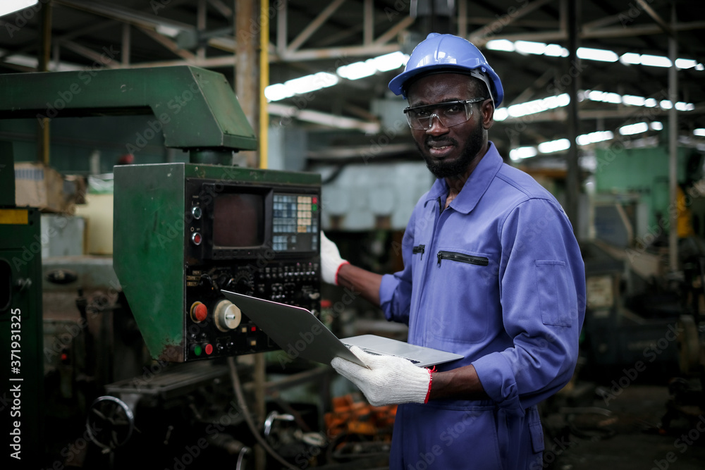 African american factory industrial engineer or worker in uniform wear safety hardhat and glasses holding laptop computer and looking at camera in manufacturing plant.maintenance and industry concept. African american factory industrial engineer or worker in uniform wear safety hardhat and glasses holding laptop computer and looking at camera in manufacturing plant.maintenance and industry concept.