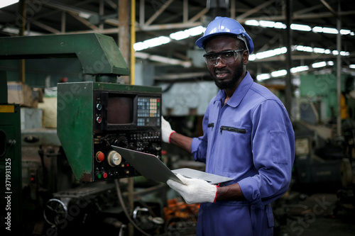 African american factory industrial engineer or worker in uniform wear safety hardhat and glasses holding laptop computer and looking at camera in manufacturing plant.maintenance and industry concept.