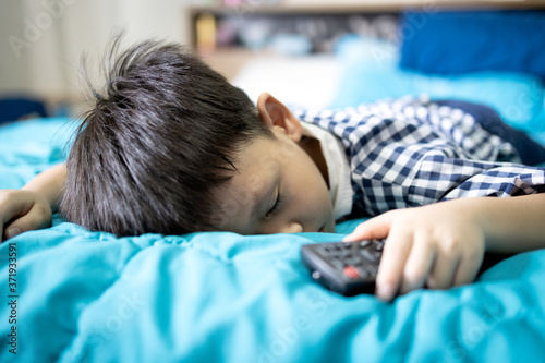 Asian kid boy is sleeping while watching cartoons online on TV with remote control in hand,little child fell asleep studying online,abnormal drowsiness,Narcolepsy disease in children,sleep disorders