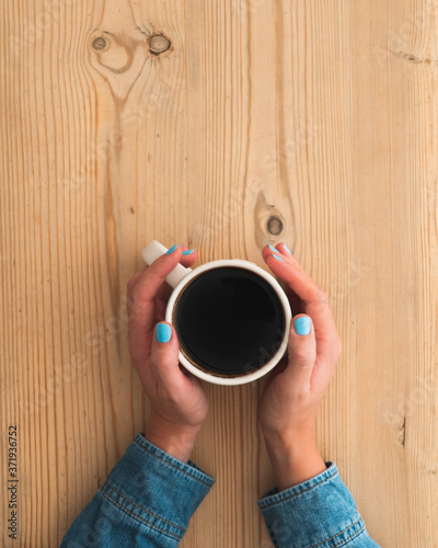 Female hands holding onto a white cup filled with black Americano coffee. Lady is wearing faded denim shirt and hands are rested on pale natural wood grain surface or table. Flat lay style image