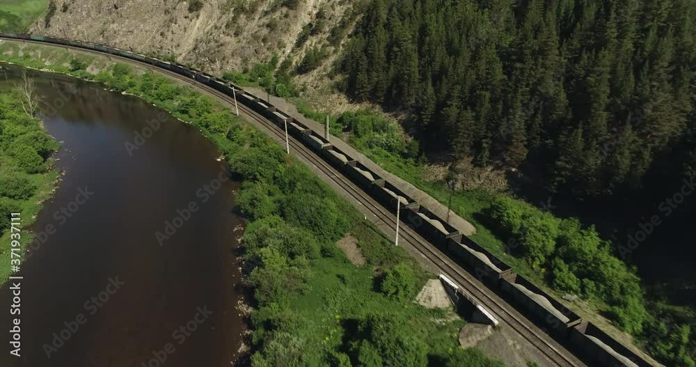 Freight long train carries with cargo carriages in a village in wild mountains landscape through a difficult part of Trans Siberian railways. Aerial drone wide view at summer day. Freight long train carries with cargo carriages in a village in wild mountains landscape through a difficult part of Trans Siberian railways. Aerial drone wide view at summer day.