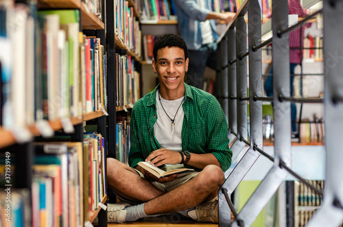 Young male student study in the library reading book while sitting near bookshelf and looking at camera 