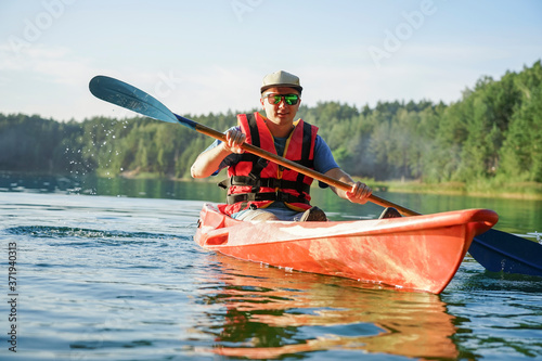 the guy is sailing on a red kayak, wearing a life jacket with glasses and a cap.