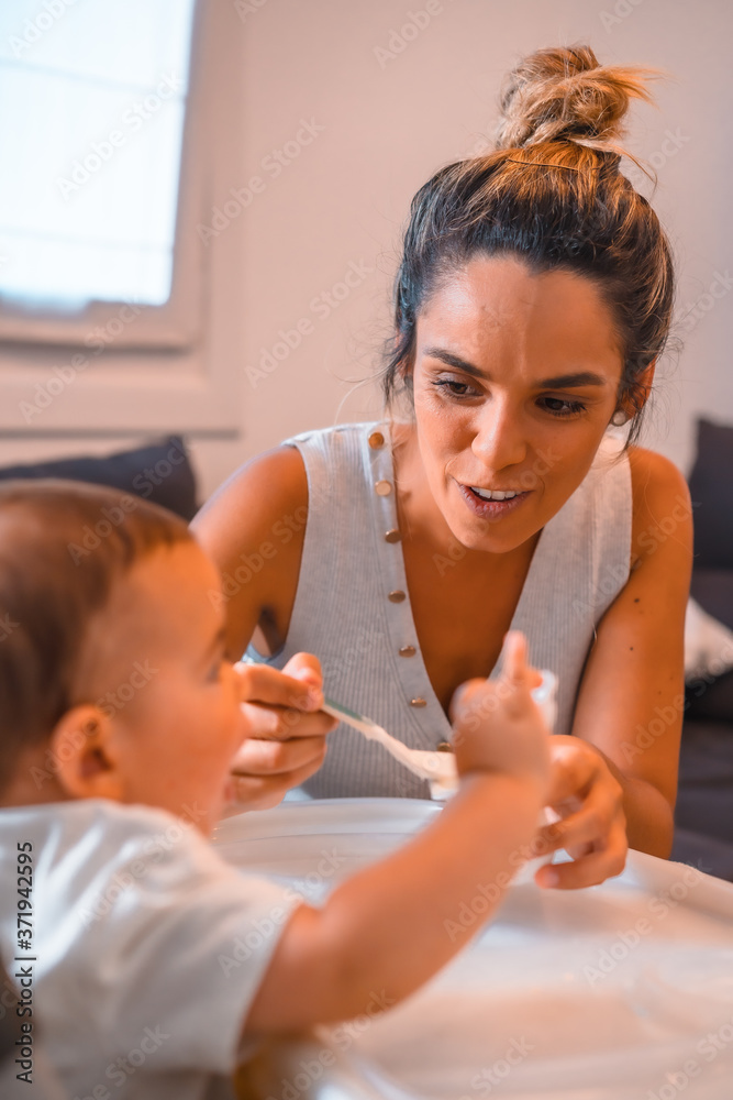 Young Caucasian mother feeding her son a yogurt while sitting in the highchair. Teleworking and caring for your child, a new normal after the coronavirus pandemic, covid-19. Young Caucasian mother feeding her son a yogurt while sitting in the highchair. Teleworking and caring for your child, a new normal after the coronavirus pandemic, covid-19.