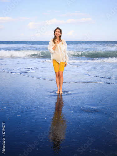 Beautiful young woman walking on black sand beach. Caucasian woman wearing yellow sportswear and white blouse. Happiness and smile. Travel lifestyle. Copy space