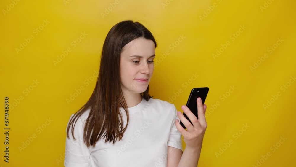 Millennial young woman use mobile phone smile while watching video on internet, social media networks user, wears t-shirt, isolated on yellow studio background. Read message feels pleased good news