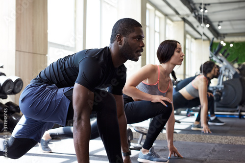 side view on sport team squatting together in gym, african guy and caucasian ladies do exercises