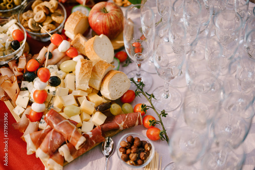 Close-up of a wooden cutting board with cold cuts and cheese and bread on a delicatessen table and empty clean champagne glasses.
