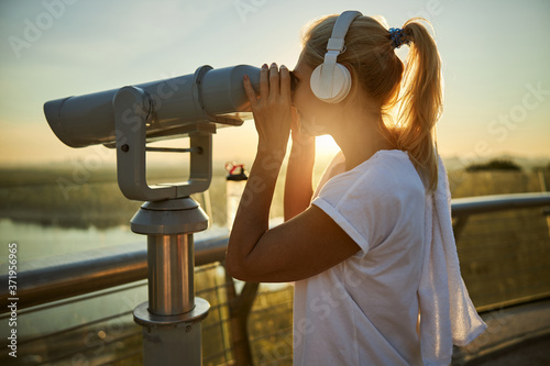 Charming woman in wireless headphones using street binoculars