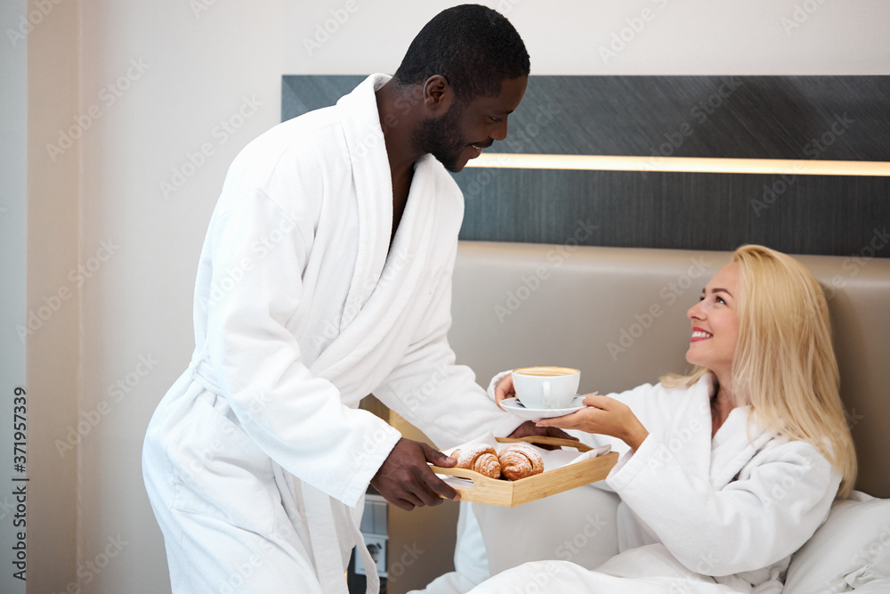 Fototapeta premium generous kind african man brought breakfast to bed to his beloved caucasian girlfriend, woman is happy to get attention by him