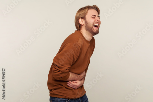 Hilarious laughter. Portrait of amused man with beard wearing sweatshirt holding belly and laughing out loud after hearing crazy anecdote, funny joke. indoor studio shot isolated on gray background