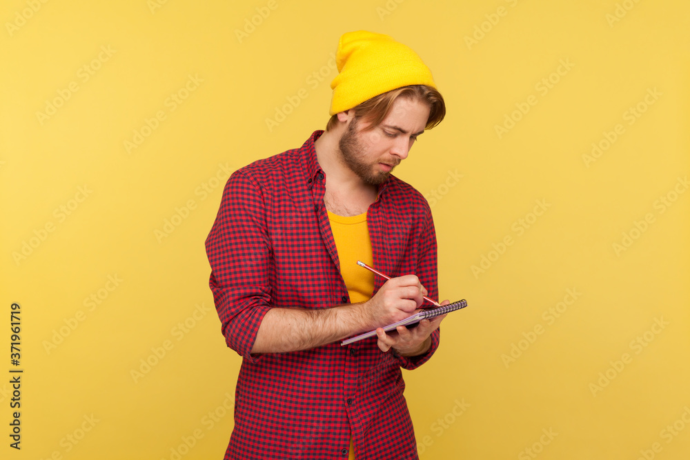 Hipster trendy guy in beanie hat looking inspired by creative idea, eager writing thought reminder to-do list into notebook, taking notes in paper, business plan. indoor studio shot isolated on yellow