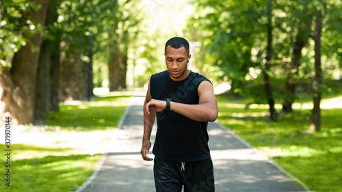 Attractive black guy looking at his fitness tracker while jogging at park