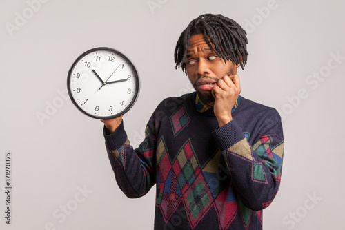 Time is out. Nervous scared african man in stylish sweatshirt anxiously looking at wall clock in his hand, frustrated with deadline, being late.