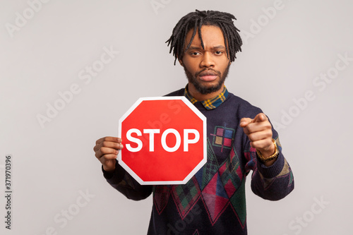 Stop! Concerned worried african guy with dreadlocks holding stop sign and pointing finger at camera with serious anxious face, give up drunk driving. Indoor studio shot isolated on gray background