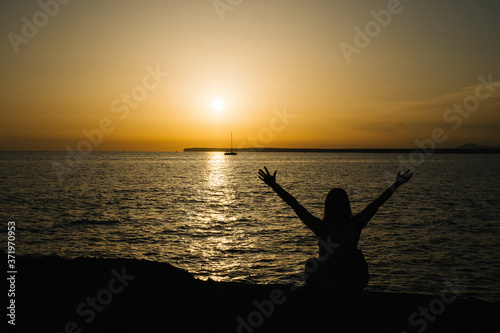 happy girl sitting with raised arms in front of the sea watching the sunset
