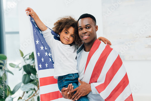 african american father and daughter wrapped in flag of america looking at camera