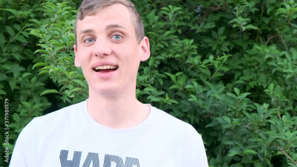 Young blond man in a white shirt smiling while looking at camera on background of green bushes, slow motion Young blond man in a white shirt smiling while looking at camera on background of green bushes, slow motion