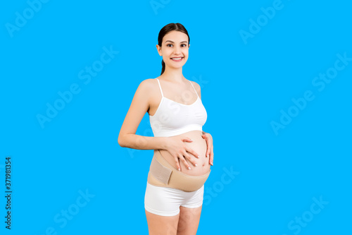 Portrait of pregnant woman in underwear wearing elastic bandage against pain in the back at blue background with copy space. Orthopedic abdominal support belt concept © sosiukin