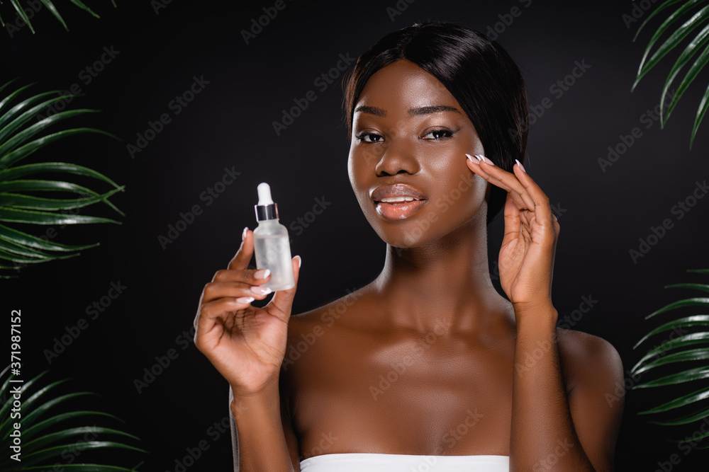 Φωτογραφία african american woman holding bottle with serum near green palm leaves isolated