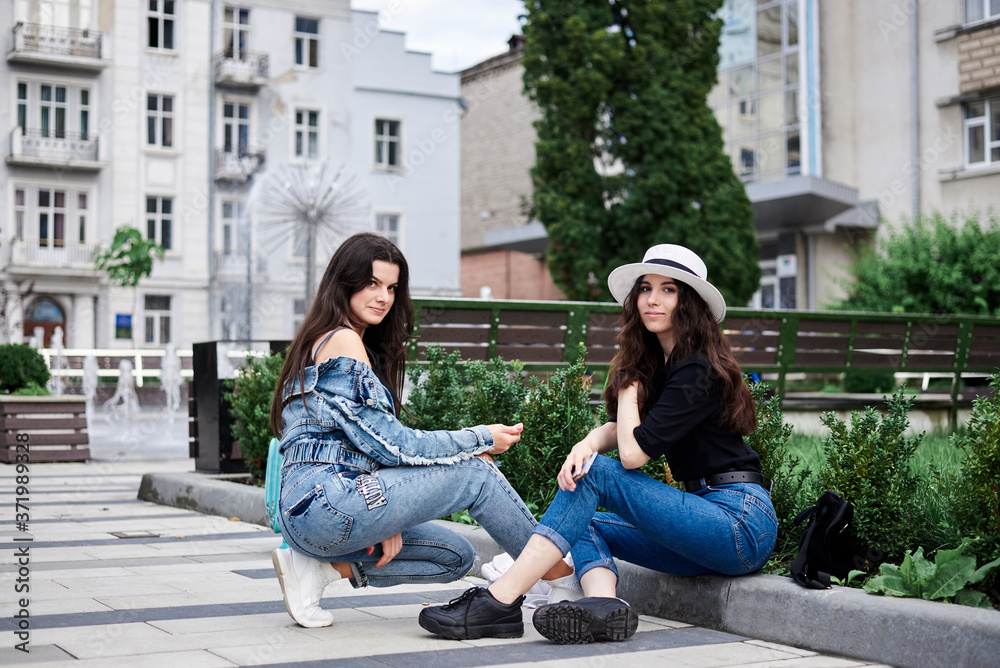 Fototapeta premium Two young brunette girls, wearing casual jeans clothes, with mint luggage, sitting on pavement border, checking phone in front of historical building in town. City sightseeing tour.Tourism lifestyle.