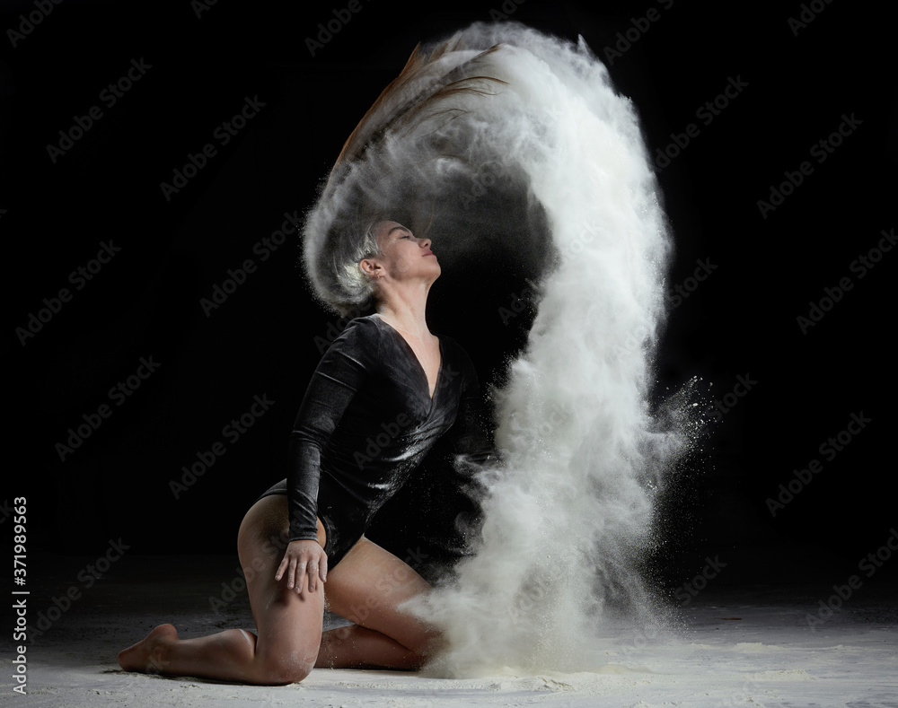 young beautiful caucasian woman with long hair is dressed in a sports black bodysuit and sits on the floor and throwing white flour up