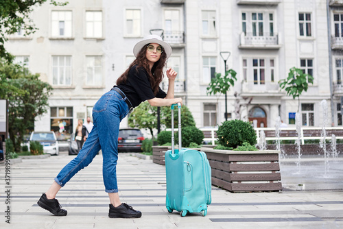 Young brunette woman, wearing black shirt, jeans and white hat with green sunglasses, holding mint luggage, walking in front of fountain in old city town in summer. Traveler in city. © Natalia