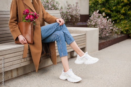 Fashionable young woman wearing beige wool coat and blue jeans. She is holding trendy tan tote bag with ranunculus flowers in hands. Street style.  photo