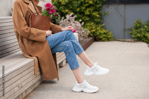 Fashionable young woman wearing beige wool coat and blue jeans. She is holding trendy tan tote bag with ranunculus flowers in hands. Street style. 