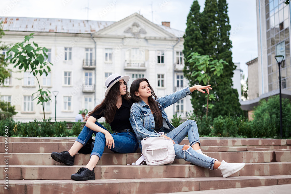 Two young brunette girls, wearing casual jeans clothes, holding small backpacks, sitting on stairway, resting, in front of historical building in town. City sightseeing tour. Traveling together.