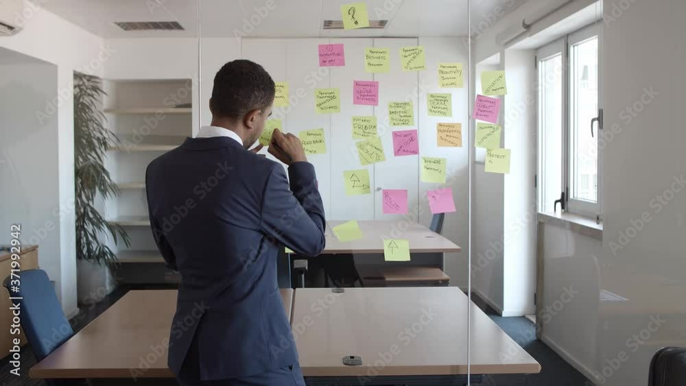 Back view of businessman writing on sticky memos on glass board in office, preparing for presentation, working on project plan. Medium shot. Management and business strategy concept Back view of businessman writing on sticky memos on glass board in office, preparing for presentation, working on project plan. Medium shot. Management and business strategy concept