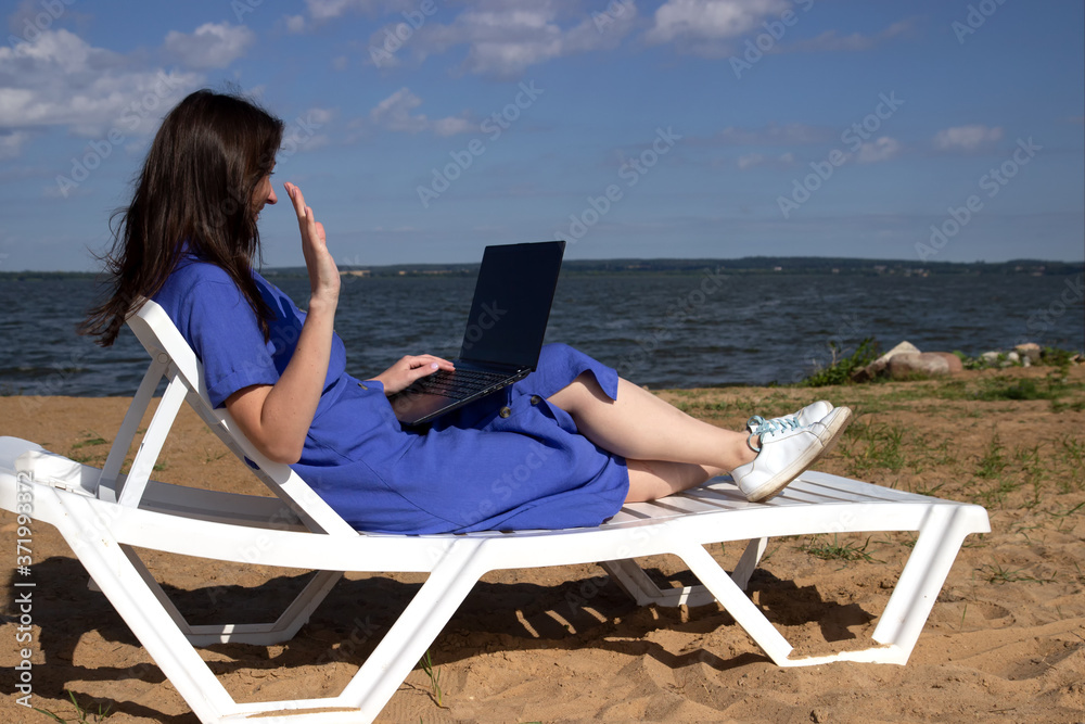 girl greeting during videocall on laptop sitting in on the beach. Freelancer and blogger participates in Videoconference
