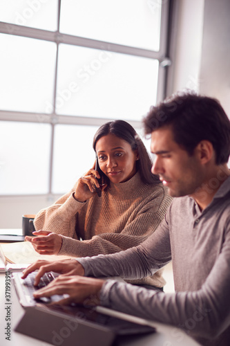 Businessman Working On Laptop At Desk Collaborating With Female Colleague Using Mobile Phone