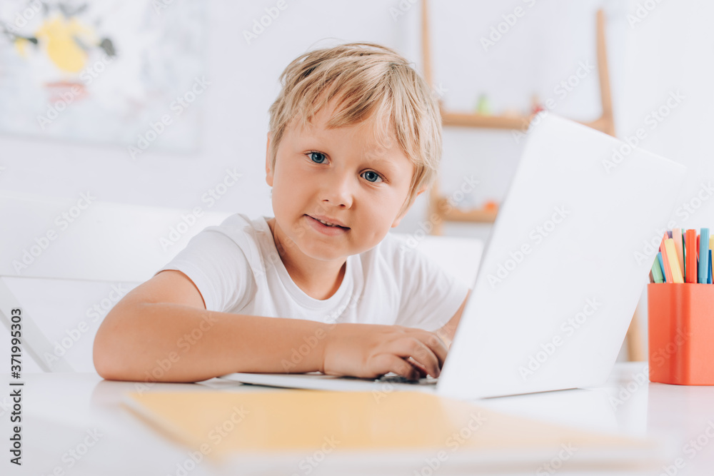 selective focus of kid in white t-shirt looking at camera while sitting at table and using laptop selective focus of kid in white t-shirt looking at camera while sitting at table and using laptop