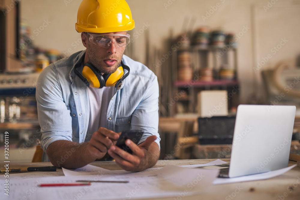 Close up of young carpenter leaned on his work desk using mobile phone Close up of young carpenter leaned on his work desk using mobile phone