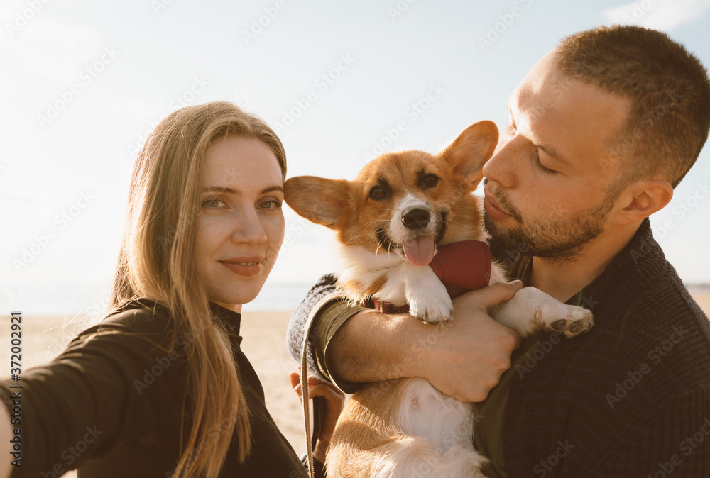 Young happy couple with dog take selfie on beach. Beautiful girl and guy and Corgi puppy are having fun. Family life, togetherness, husband and wife relax together in summer by sea