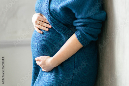 Close-up shot of belly of a 30 weeks pregnant woman wearing a navy blue bodycon dress in a light room. Pregnancy planning and preparation for childbirth