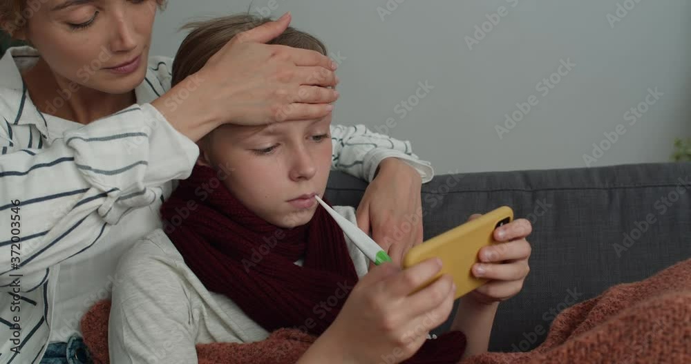 Close up of pretty woman touching her teen son forehead and carrying of him while they sitting on sofa. Sick kid measuring temperature and playing online game on smartphone near mom