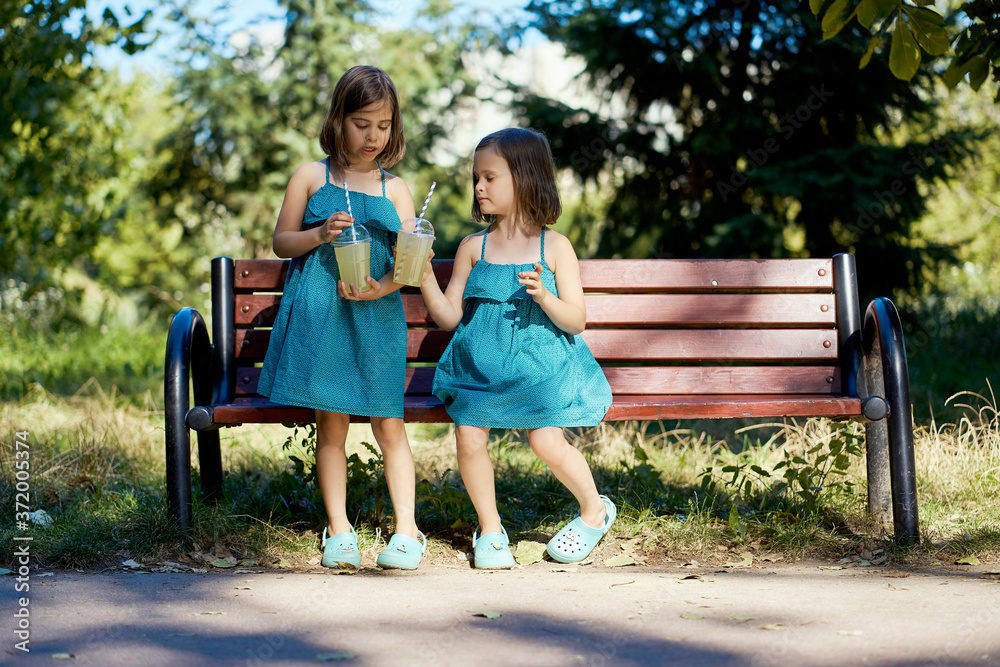 two cute little girls in dresses sit on a bench and drink lemonade in the summer two cute little girls in dresses sit on a bench and drink lemonade in the summer