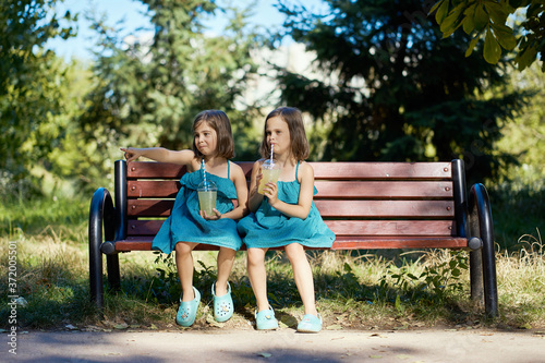 Obraz na płótnie two cute little girls in dresses sit on a bench and drink lemonade in the summer w salonie