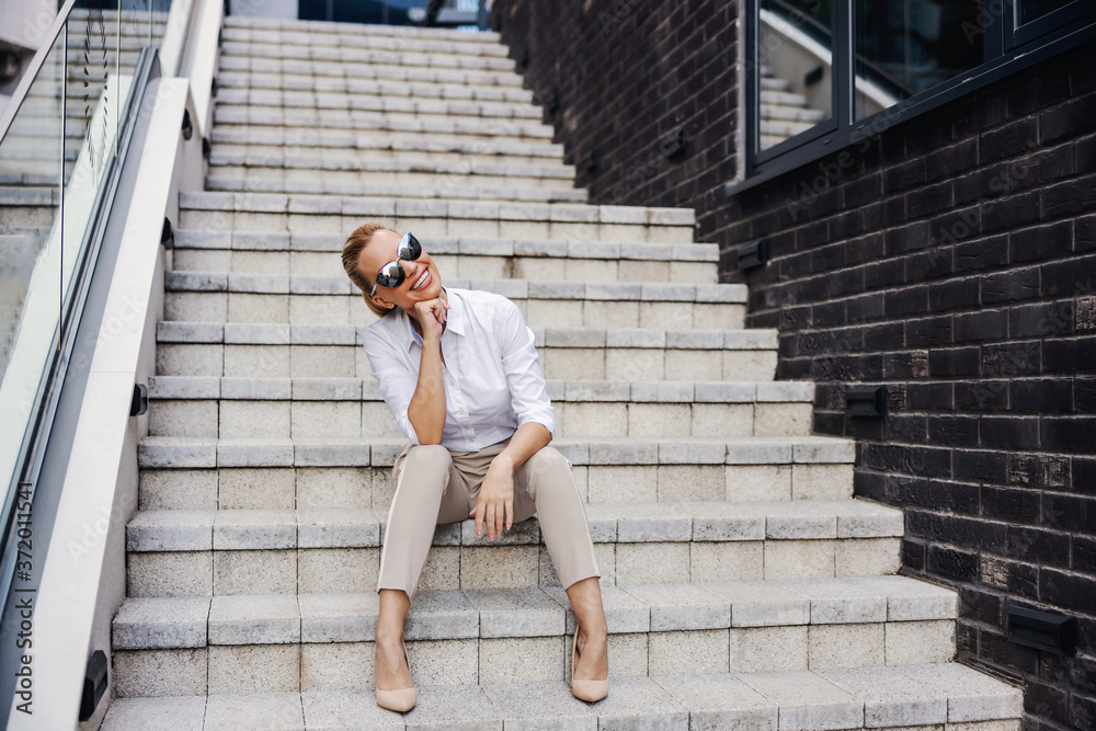 Beautiful smiling fashionable businesswoman sitting on the stairs in front of business center and posing.