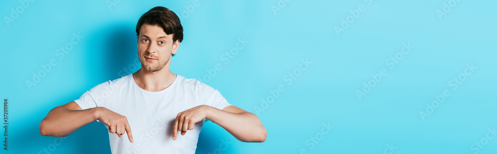 Panoramic shot of young man in white t-shirt pointing with fingers on blue background Panoramic shot of young man in white t-shirt pointing with fingers on blue background