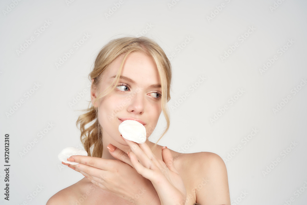 A blonde with bare shoulders holds cotton pads near her face skin care light background A blonde with bare shoulders holds cotton pads near her face skin care light background