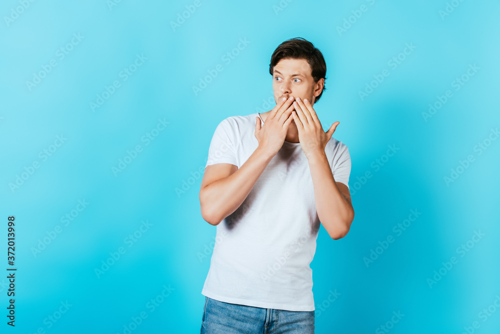 Surprised man in white t-shirt covering mouth with hands on blue background Surprised man in white t-shirt covering mouth with hands on blue background
