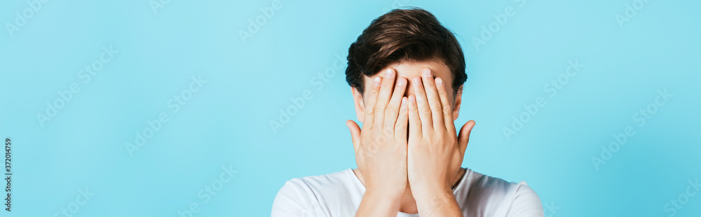 Panoramic shot of man covering face with hands on blue background Panoramic shot of man covering face with hands on blue background