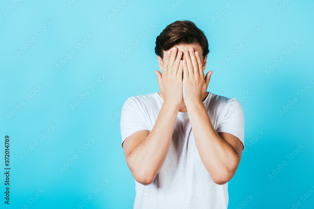 Frightened man in white t-shirt covering face with hands on blue background Frightened man in white t-shirt covering face with hands on blue background